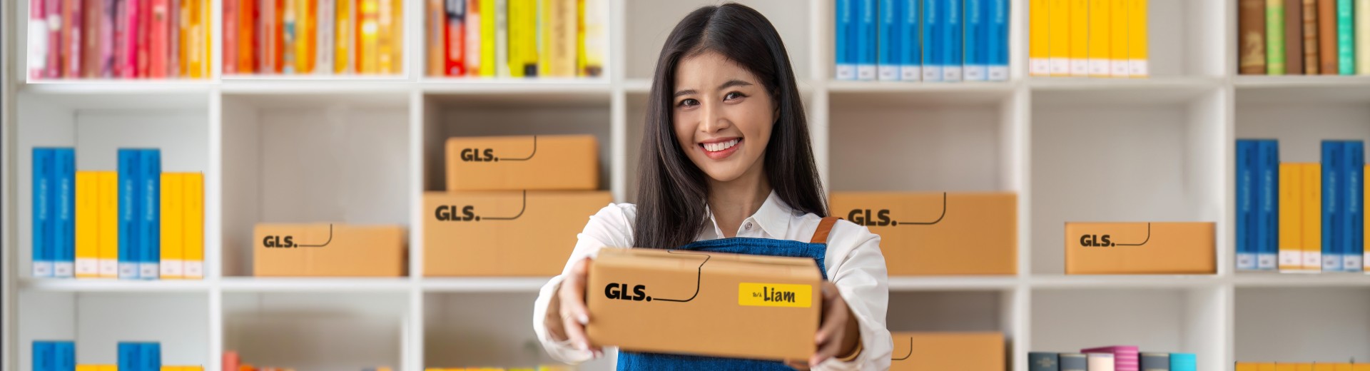 Woman handing out a GLS parcel. She stands at a desk in front of shelves field with colourful books.