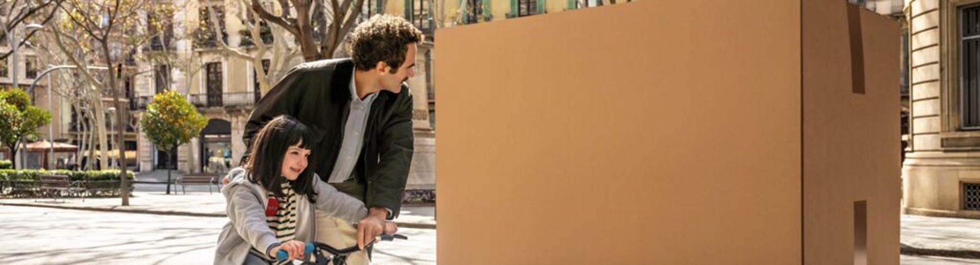 Ein Vater betrachtet ein großes Paket, während er seiner Tochter das Fahrradfahren beibringt.